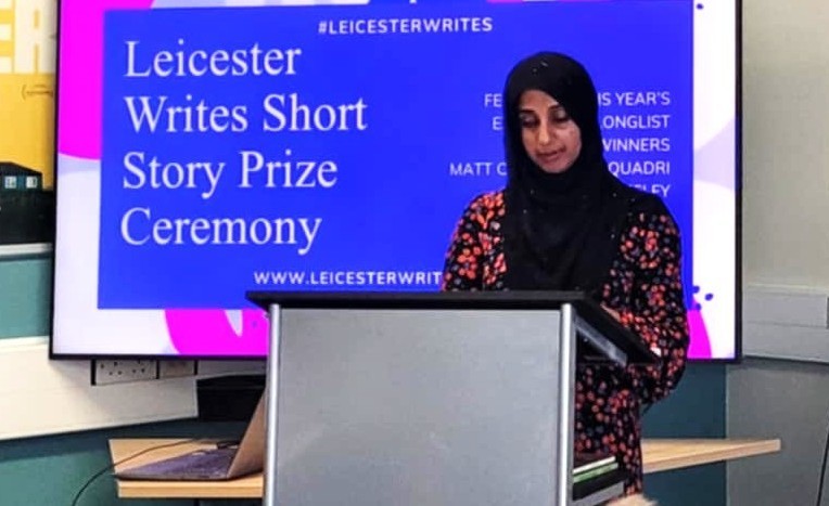 Woman in a hijab, publisher Farhana Sheikh, stands in front of a sign for the Leicester Writes Short Story Prize Ceremony.